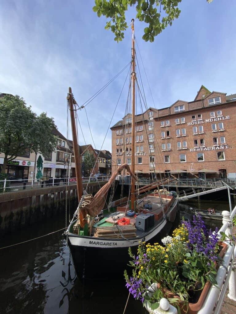 Traditionelles Segelschiff "Margareta" im Wasser, umgeben von Blumen und Stadtgebäuden.