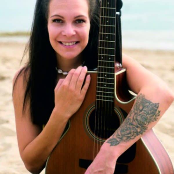Lea mit Gitarre am Strand, lächelnd, bei schönem Wetter.